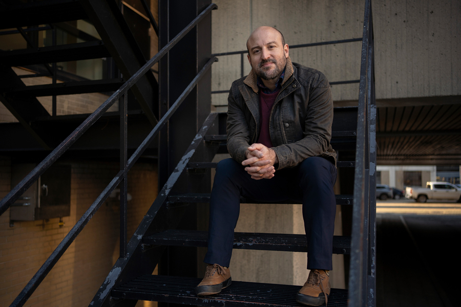 Otto Cordero sits on an outdoor black metal staircase with buildings and car in background.