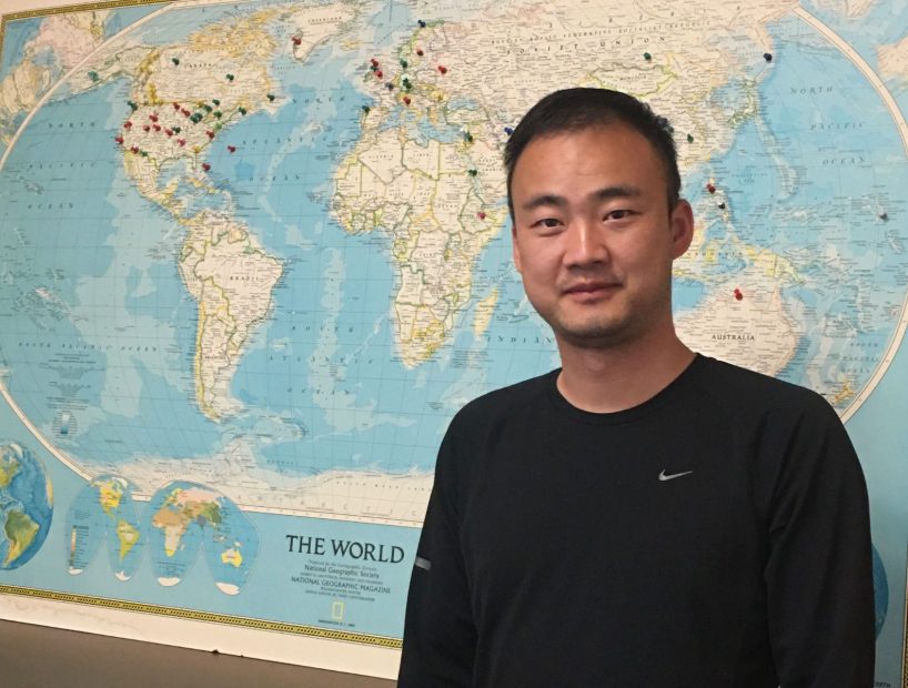 Headshot of Wenzhe Jiao standing in front of a map of the world.